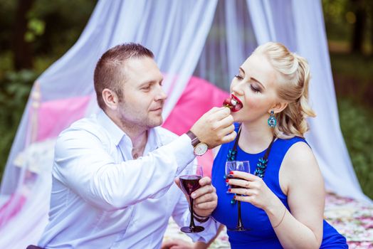 Man and woman sitting on the bed in the lawn with glasses of wine and woman eating strawberries from a man's hand in Lviv, Ukraine.