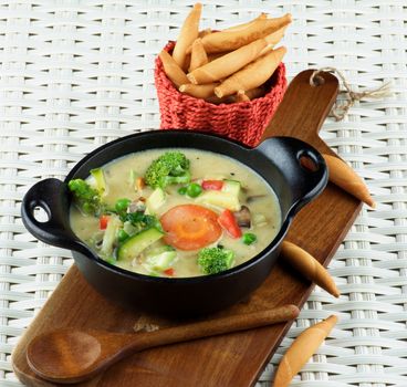 Homemade Vegetables Creamy Soup in Black Stewpot with Wooden Spoon and Bread Sticks on Wooden Cutting Board closeup on Wicker background