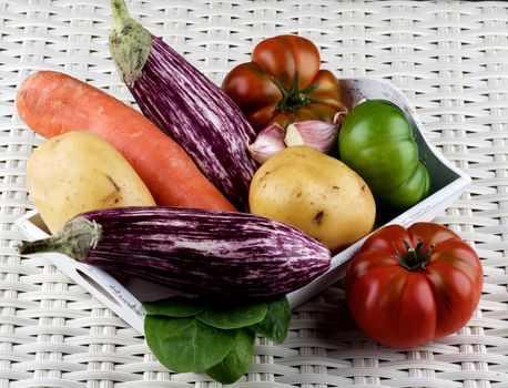 Arrangement of Fresh Raw Vegetables with Striped Eggplants, Potato, Carrot, Green and Red Tomatoes, and Garlic on Wooden Tray closeup on Wicker background