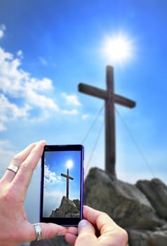 Photographing and taking photo of wooden cross on peak by smart phone. View over smart phone screen during photographing of landscape shot.