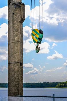 Yellow-green hook of a construction crane hanging on a rope on a background of clouds on a lake dam