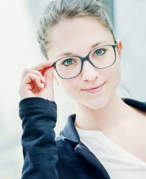 Blonde girl standing outdoor. She holds glasses on face. She is looking to the camera.