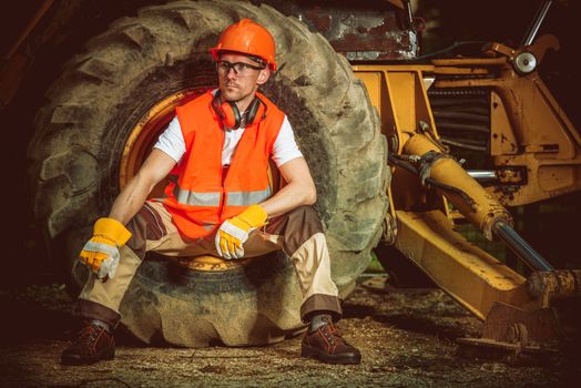Caucasian Construction Worker While Seating Inside Large Dozer Wheel. Construction Site Concept.