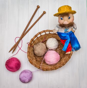 From above the colored balls of yarn in wicker basket with needles and traditional toy of man on white wooden board in Ukraine