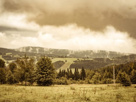 Aged nostalgic weathered vintage retro photo of cloudy misty landscape. mountain meadow and forests, both deciduous broadleaved and needle coniferous trees, electric overhead power line and horizon covered in clouds and mist, Czech republic, central Europe, Orlicke hory