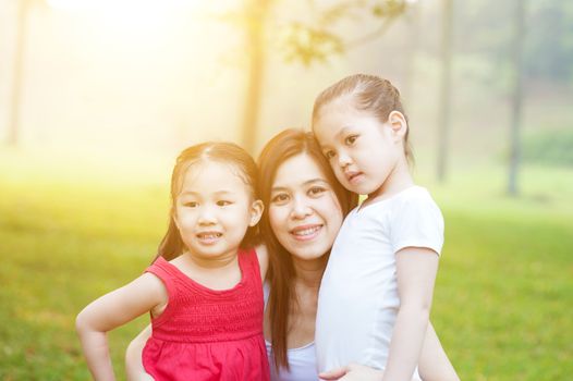 Pretty Asian woman looking at camera with smile while holding her preschool daughters in arms, little girl hugging her mother tenderly, morning with sun flare.