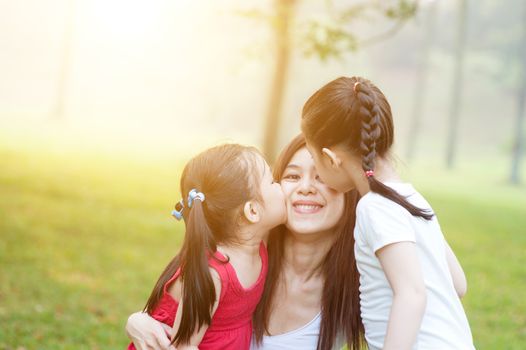 Lifestyle portrait of Asian mother and daughters kissing in the park, Family outdoor fun, morning with sun flare.