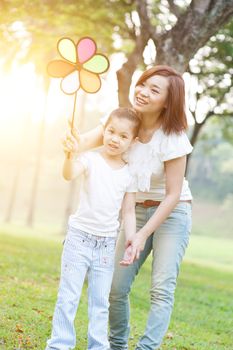 Happy Asian mother and daughter playing windmill in the park. Family outdoor fun, morning with sun flare.
