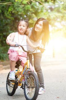 Asian little girl learning ride on bicycle with help of mother in the park, family outdoor fun activity.