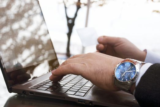 The rear view of male hands holding a credit card by typing on laptop computers while sitting across the image.