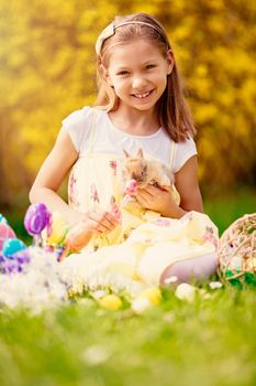 Beautiful smiling little girl holding cute bunny and sitting on the grass in spring holidays. Looking at camera.