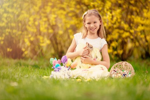 Beautiful smiling little girl holding cute bunny and sitting on the grass with Easter eggs in spring holidays. Looking at camera. Copy space.