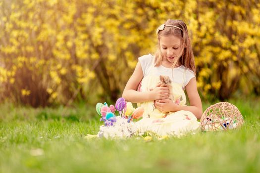 Beautiful smiling little girl holding cute bunny and sitting on the grass with Easter eggs in spring holidays. Copy space.