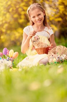 Beautiful smiling little girl holding cute bunny in the basket and sitting on the grass with Easter eggs in spring holidays. Looking at camera. Copy space.
