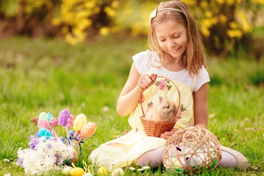 Beautiful smiling little girl holding cute bunny in the basket and sitting on the grass with Easter eggs in spring holidays. 