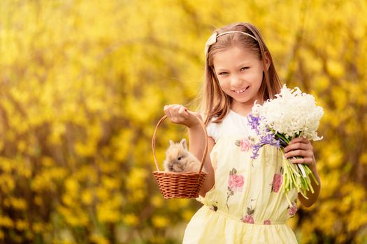 Beautiful smiling little girl holding cute bunny in the basket and bouquet white flowers in spring holidays. Looking at camera. Copy space.