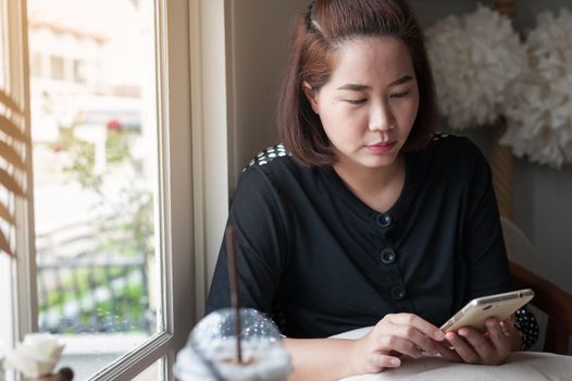 Young Asian woman using smartphone for browsing internet, personal chat, and social media in coffee shop on weekend. Vacation lifestyle in cafe