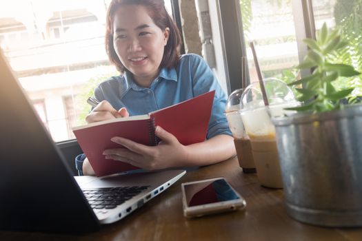 Young Asian hipster woman writing on notebook while working with laptop computer in coffee shop. Freelance business starup activity concept