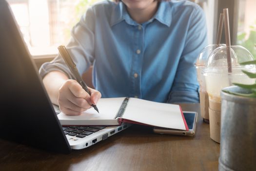 Young Asian hipster woman writing on notebook by right hand while working with laptop computer in coffee shop. Freelance business starup activity concept