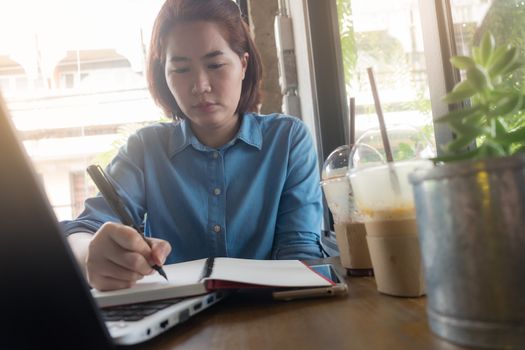 Young Asian hipster woman writing journal on notebook with serious emotion in coffee shop. Freelance business starup activity concept