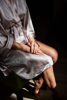 A girl in a silk robe sits on a chair and crosses her hands on her knees.