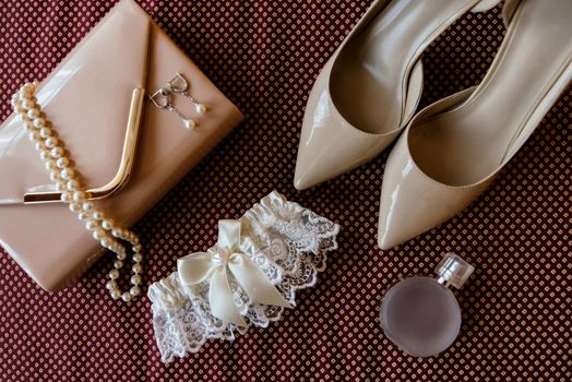 Bride's shoes, garter, perfume, necklace, earrings and clutch are on burgundy cloth surface