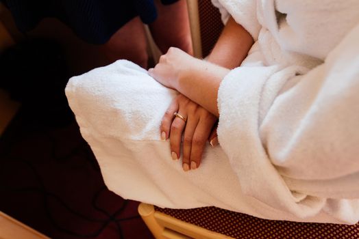 A girl in a white robe sits on a chair and crosses her hands on her knees