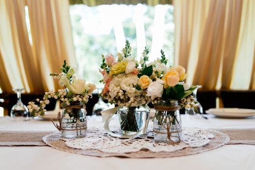 Wedding bouquets of yellow, white and pink roses are in vases on dinner table