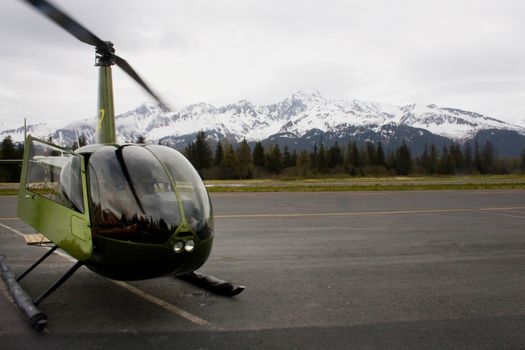 A small green helicopter preparing to take off in Seward Alaska with mountains in background. 