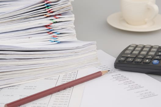 Brown pencil and calculator on finance account have blur pile overload document of report and receipt with colorful paperclip and cup of coffee as background.