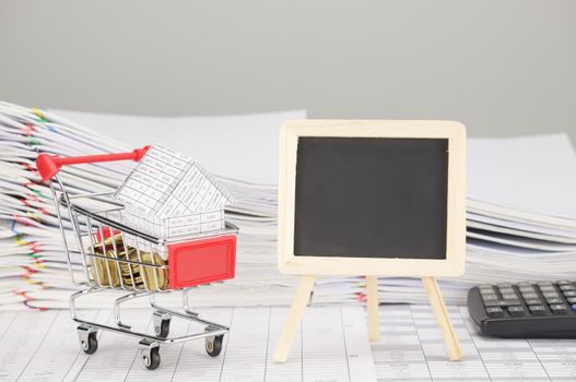 House in shopping cart with gold coins and empty blackboard on finance account have blur pile overload document of report and receipt with colorful paperclip and calculator as background.