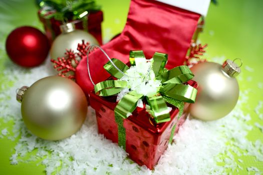 Holiday Present - Red Present Box with Green Glossy Bow. Some Christmas Ornaments and Fake Snow. Green Background