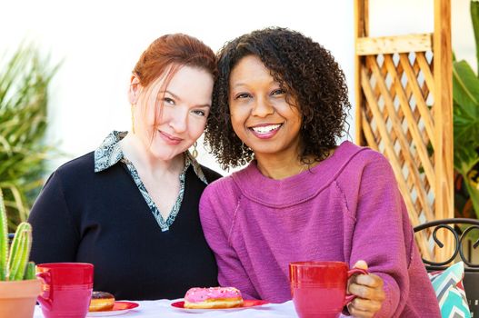 Happy adult mixed lesbian couple sitting together at table with coffee and donuts