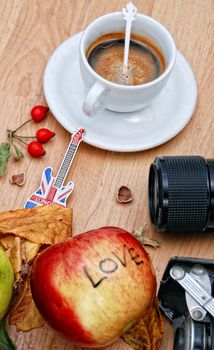 White cup of espresso, souvenir guitar from london, camera lens, rose hips and apple with text love