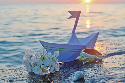 Paper boat, seashells and a sprig of cherry blossoms on a large stone on the seashore