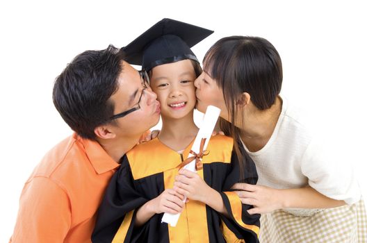 Asian kindergarten child in graduation gown and mortarboard kissed by her parent during graduation