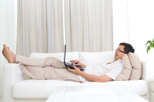 Online apps concept. Guy using laptop. Handsome asian man relaxed and lying on sofa indoor.