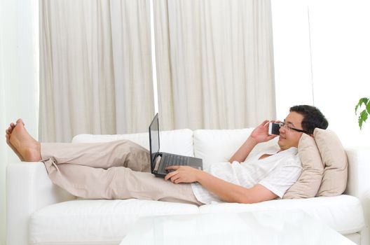 Smartphone mobile apps concept. Guy using smart phone and laptop to perform internet banking. Handsome asian man relaxed and lying on sofa indoor.