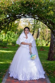 A stylish bride with a bouquet in her hands is standing under a green arch