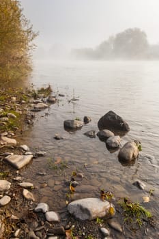 foggy shore of the river in autumn with small boulders in the foreground