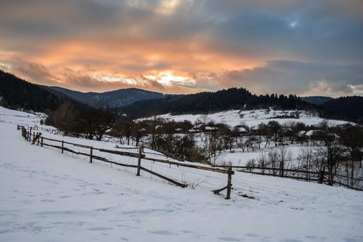 road to the village covered with snow in the mountains under a red winter sunset