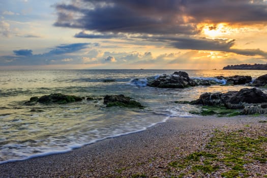 rays of the morning sun behind the clouds illuminate sea and coastal sandy beach with small rocks