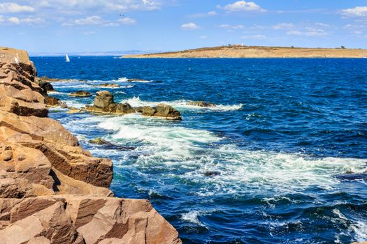 Sea waves break on the huge rocks on the shore. on the other side of of seascape can be seen bald island with hill and buildings. small sailboat sails in the distance between the shore and the island