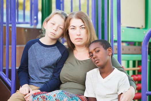 Sad mother sitting with pair of unhappy male children at playground