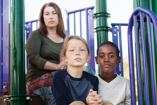 Two male children sitting on playground set with mother behind them