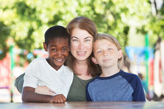 Mother hugging and sitting in between adopted child and son at table in park