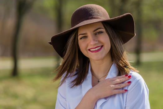 Portrait of a smiling young woman, holding her hand on her shoulder and wearing a hat in a park during spring. Woman bowed her head head to the left side of frame. Medium shot. Shallow depth of field.