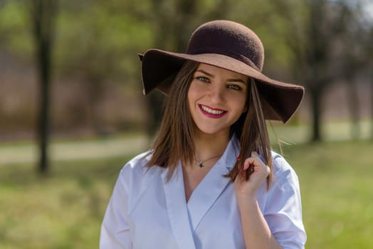 Portrait of a smiling young woman wearing a hat in a park during spring. Woman bowed her head head to the right side of frame. Medium shot. Shallow depth of field.
