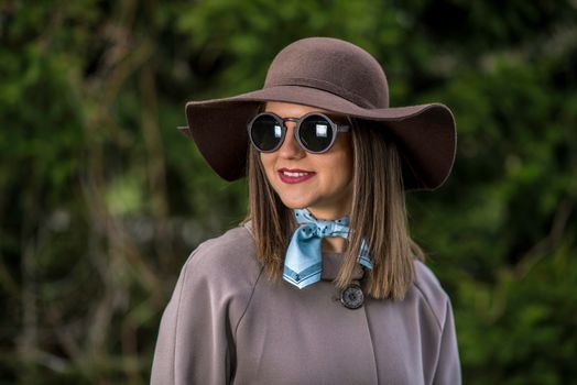 Young woman in hat, raincoat and sunglasses is posing in the park during a sunny spring day. Shallow depth of field.