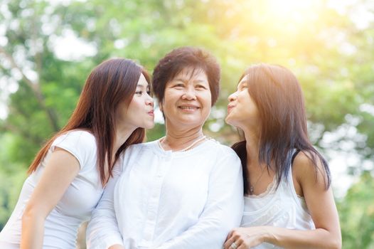 Portrait of happy Asian daughters kissing elderly mother, senior adult woman and grown child. Outdoors family at nature park with beautiful sun flare.
